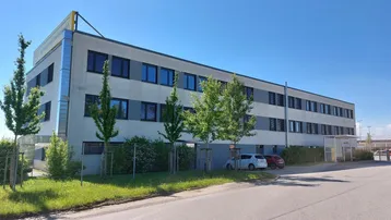 Modernes Geschäftsgebäude mit grauer Fassade, vielen Fenstern und gepflegter Außenanlage unter blauem Himmel.