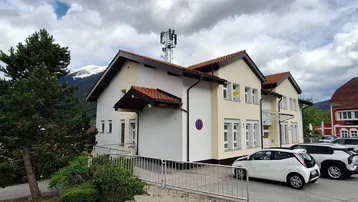 Gebäudeansicht mit Bergen im Hintergrund und parkenden Autos auf der Straße.