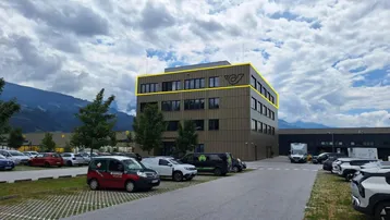 Modernes Geschäftsgebäude mit großem Parkplatz und Bergkulisse unter bewölktem Himmel.