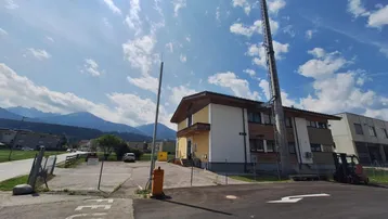 Gebäude mit gelber Fassade, Holzelementen und Satteldach, umgeben von einem Parkplatz und Bergen im Hintergrund.