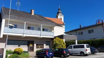 Gebäudeansicht mit Balkon, Parkplätzen und einer Kirche im Hintergrund unter blauem Himmel.
