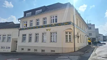 Historisches Gebäude mit heller Fassade, vielen Fenstern und dekorativen Elementen an einer Straßenecke.