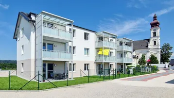 Modernes Mehrfamilienhaus mit Balkonen und Terrassen in ländlicher Umgebung neben einer Kirche.