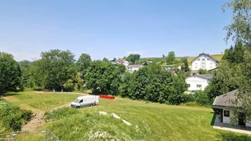 Weitläufiger Blick über grüne Felder und eine ländliche Siedlung unter blauem Himmel.