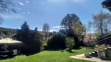 Großer, gepflegter Garten mit Rasenfläche, Bäumen und einem gemütlichen Sitzbereich unter einem Sonnenschirm.