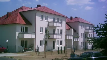Zwei neuwertige Mehrfamilienhäuser mit roten Ziegeldächern und mehreren Balkonen, umgeben von Grünflächen unter blauem Himmel.
