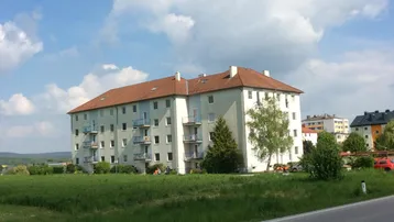 Mehrfamilienhaus mit hellblauer Fassade, rotem Ziegeldach und Balkonen, umgeben von grünen Feldern unter blauem Himmel.