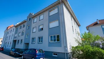 Mehrstöckiges Wohngebäude mit hellgrauer und blauer Fassade unter klarem blauem Himmel und parkenden Autos.