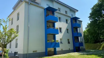 Modernes Mehrfamilienhaus mit hellen Fassaden und markanten blauen Balkonen, umgeben von einer gepflegten Grünfläche.