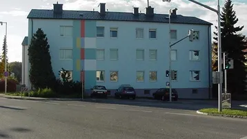 Mehrfamilienhaus mit hellblauer Fassade und farbigen Streifen an einer belebten Straße mit Parkplätzen.