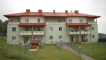 Mehrfamilienhaus mit grüner Fassade, roten Dächern, Balkonen und einem gepflegten Garten mit Spielbereich.