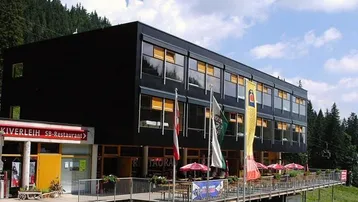 Modernes Gebäude mit schwarzer Fassade, großen Fenstern und einer Außenterrasse in bergiger Umgebung.
