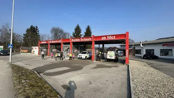 Außenansicht einer modernen SB-Waschanlage mit mehreren Waschboxen und Kunden bei der Autopflege an einem sonnigen Tag.