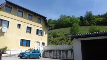 Gelbes Wohnhaus mit einem Auto davor, einer Garage und grünen Hügeln im Hintergrund unter blauem Himmel.