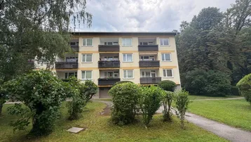 Mehrfamilienhaus mit mehreren Balkonen, umgeben von Grünflächen und Bäumen unter bewölktem Himmel.