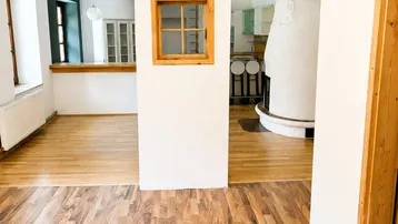 Geräumiger Wohnbereich mit Holzboden, dekorativem Kachelofen und Durchgang zur Küche.