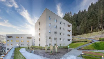 Neubauwohnungen mit moderner Architektur und Balkonen, eingebettet in eine grüne Berglandschaft unter blauem Himmel.