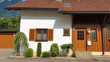 Traditionelles Haus mit weißer Fassade, Holzverkleidung und Satteldach vor einer Bergkulisse.