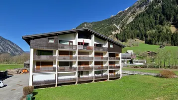 Modernes Apartmentgebäude in alpiner Landschaft mit Balkonen und Bergblick unter blauem Himmel.