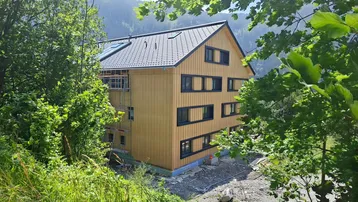 Neubau mit Holzfassade und dunklem Dach, umgeben von grüner Natur und Bergen.