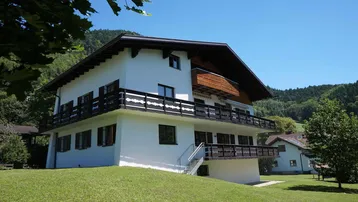 Weißes Haus mit traditionellen Holzdetails und Balkonen, umgeben von grüner Natur und Bergen.