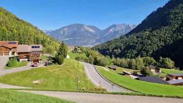 Panoramablick auf ein grünes Tal mit Häusern, einer Straße und majestätischen Bergen im Hintergrund unter blauem Himmel.