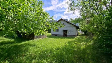 Kleines, älteres Haus umgeben von viel Grün und Bäumen, mit Bergblick im Hintergrund.