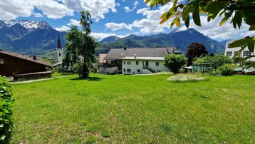 Weitläufige Grünfläche mit Blick auf die umliegende Berglandschaft und ein traditionelles Gebäude mit Kirchturm.