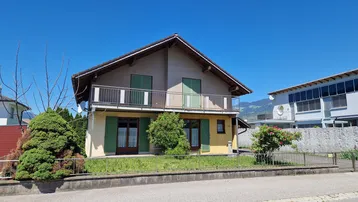 Traditionelles Haus mit Balkon, grünen Fensterläden und einem kleinen Vorgarten unter blauem Himmel.