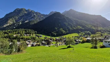 Panoramablick auf ein malerisches Dorf in den Alpen, umgeben von grünen Wiesen und majestätischen Bergen unter strahlend blauem Himmel.