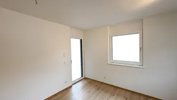 Leeres Zimmer mit hellem Holzfußboden und Zugang zu einem französischen Balkon.