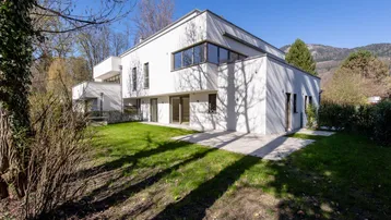 Moderne weiße Architektur mit Flachdach und großen Fenstern, umgeben von einem grünen Garten.