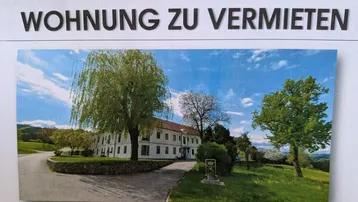 Historisches Gebäude mit weitläufiger Grünfläche und altem Baumbestand unter blauem Himmel.