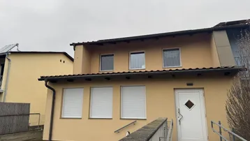 Fassade eines Gebäudes mit gelbem Putz, weißen Rollläden und einer Rampe am Eingangsbereich.