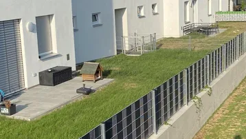 Moderne weiße Gebäude mit gepflegtem Rasen, Terrasse und einem Zaun mit Solarpaneelen.