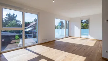 Helles, leeres Wohnzimmer mit großen Fenstern, Holzboden und Zugang zu einem Balkon.