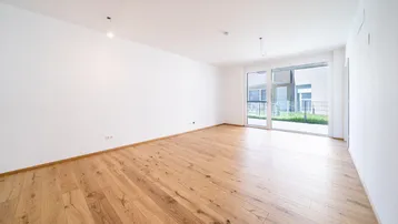 Helles, leeres Zimmer mit Holzboden und Zugang zu einem Außenbereich durch eine große Glasschiebetür.