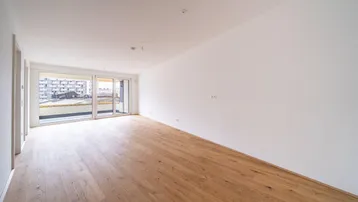 Helles, leeres Wohnzimmer mit Holzboden und Zugang zu einem Balkon, bietet viel natürliches Licht.