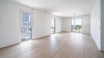 Helles, leeres Zimmer mit hellem Holzboden und großen Fenstern, die viel Tageslicht hereinlassen.