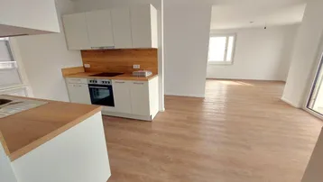 Heller offener Wohnbereich mit integrierter Küche, modernem Laminatboden und Zugang zum Balkon.