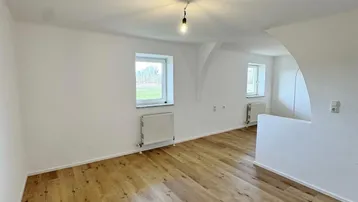 Heller, leerer Raum mit Holzboden, weißen Wänden und Fenstern, die viel Tageslicht hereinlassen.