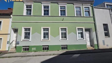 Grünes Mehrfamilienhaus mit mehreren Fenstern und zwei Eingangstüren an einer Straße gelegen.