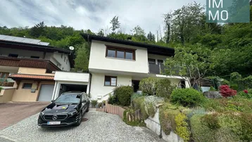 Hausfassade mit Auffahrt und Garage, umgeben von grüner Vegetation und Bäumen.