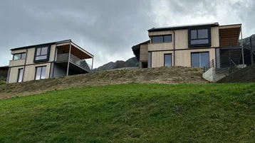 Moderne Holzhäuser auf einem grünen Hügel mit Bergblick unter bewölktem Himmel.