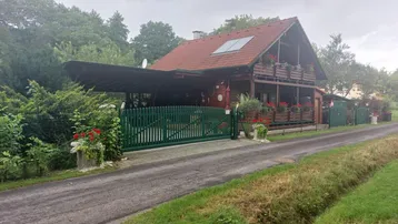 Rustikales Holzhaus mit rotem Ziegeldach, grünem Zaun und überdachtem Carport an einer ländlichen Straße.