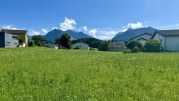 Grüne Wiese mit Häusern im Hintergrund und majestätischem Bergblick unter blauem Himmel.