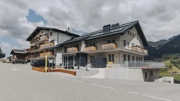 Modernes Gebäudeensemble mit traditionellen Holzbalkonen und Photovoltaikanlage in alpiner Umgebung, umgeben von grünen Hügeln.
