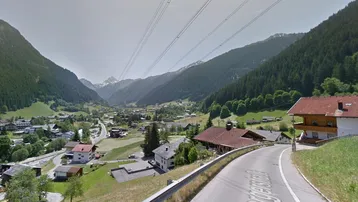 Panoramablick auf ein alpines Tal mit Häusern, Bergen und einer Straße, die sich durch die Landschaft schlängelt.