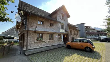 Traditionelles Gebäude mit Holzfassade und Schindeldach, umgeben von einem Parkplatz unter strahlendem Himmel.