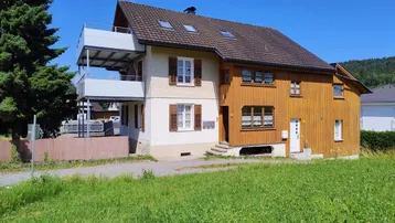 Freistehendes Haus mit Holzelementen und einem Balkon in ländlicher Umgebung unter blauem Himmel.